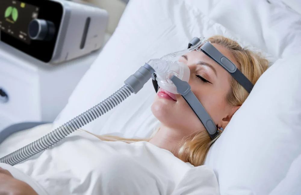 "Kvinne sover med en CPAP-maske for behandling av søvnapné, koblet til en maskin, i en seng med hvitt sengetøy."