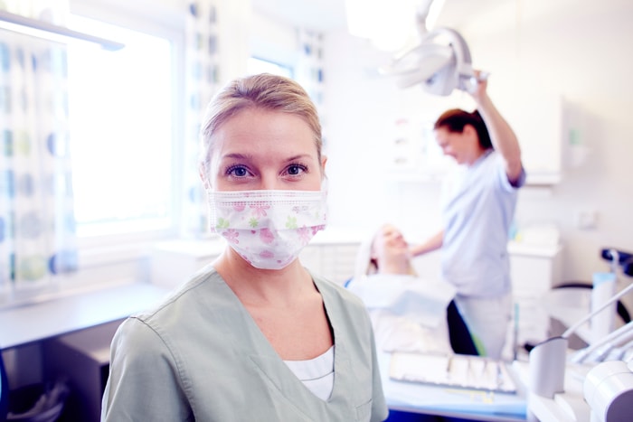 "Tannpleier med maske smiler i et lyst tannlegekontor, mens en kollega behandler en pasient i bakgrunnen."