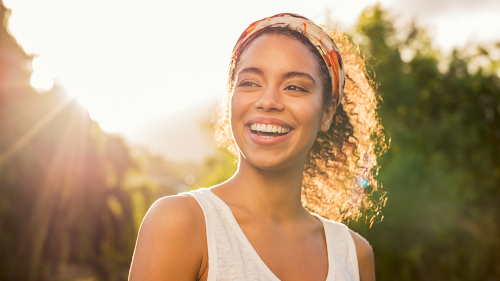 Ung kvinne smiler bredt i sollys utendørs, med naturlig og friskt uttrykk. Bildet formidler selvtillit og livsglede – et harmonisk smil i fokus.