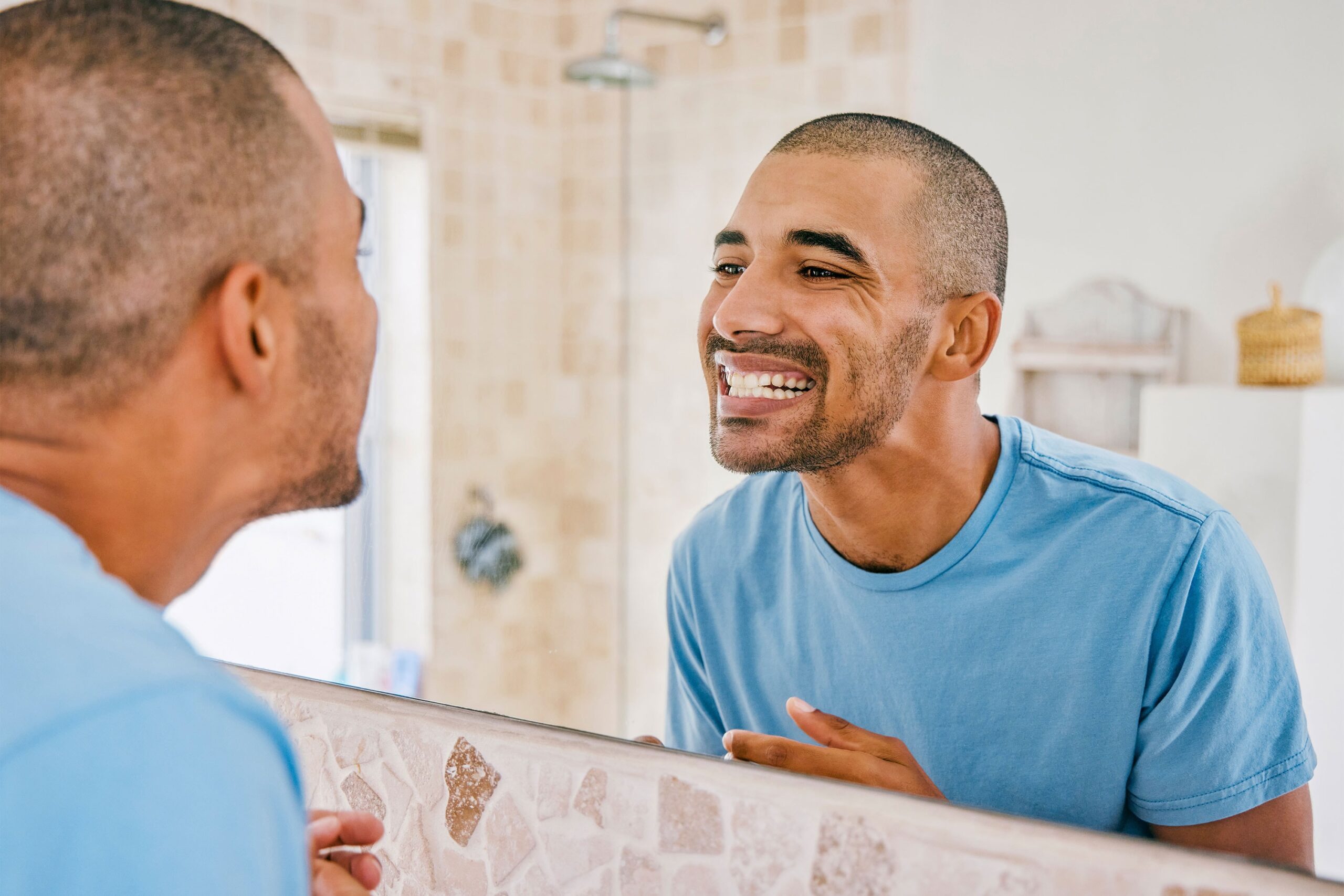 Mann smiler og sjekker tennene sine i speilet på badet, med fokus på oral helse og selvpleie i et lyst miljø."