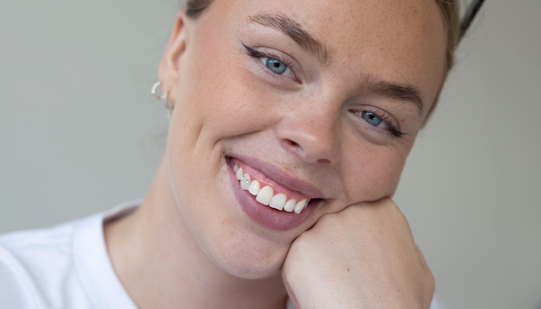 En kvinne smiler varmt med hodet lent mot hånden, og viser et smykke på en tann mens hun ser rett mot kameraet.
