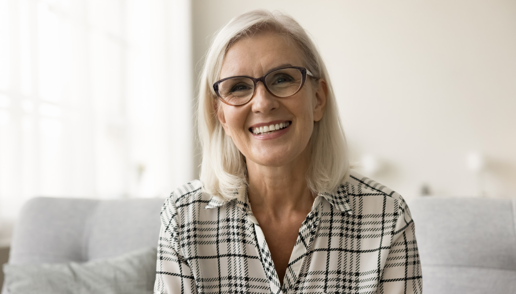 "Eldre kvinne med briller og lyst hår smiler vennlig mens hun sitter i en lys og koselig stue."