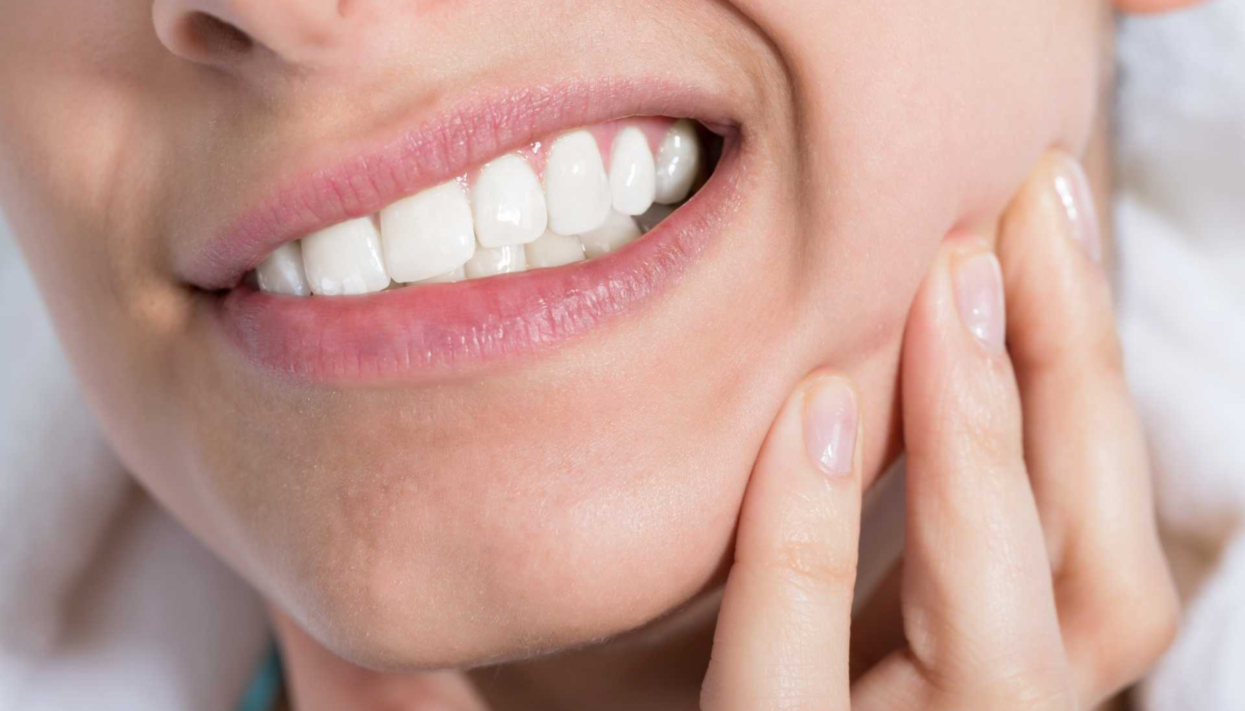  Nærbilde av en person som smiler med synlige tenner mens hånden trykker mot kinnet, som ved smerte i kjeveområdet.