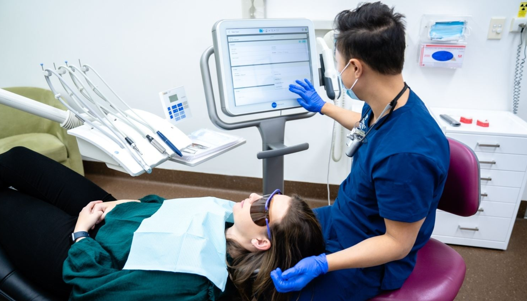 Tannlege med blå hansker og munnbind viser pasient informasjon på skjerm under tannbehandling i klinikk.