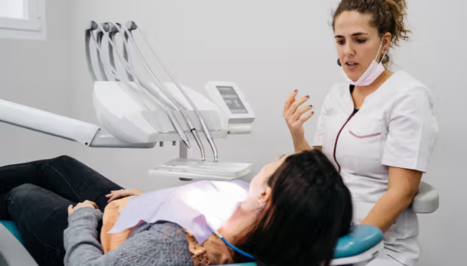Tannlege i samtale med pasient som ligger i tannlegestolen, under en tannbehandlingslampe i klinikkmiljø.