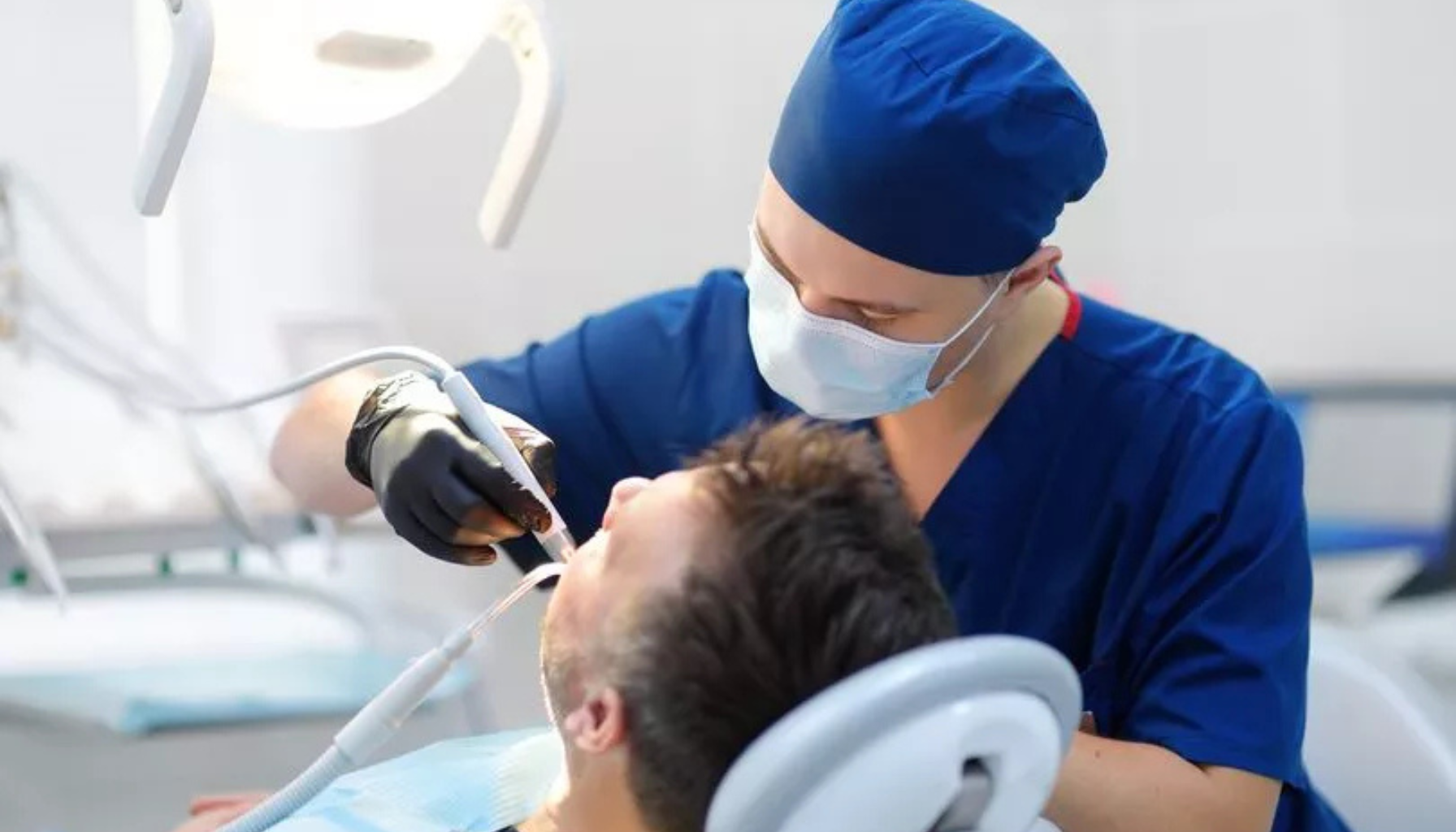 Tannlege i blå uniform og munnbind behandler pasient med moderne utstyr under sterkt lys i et klinisk og profesjonelt tannlegekontor.